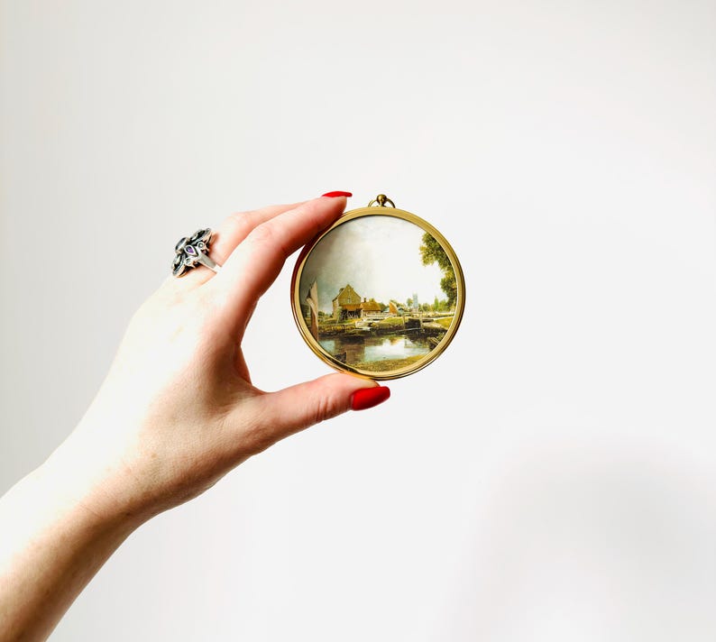 May include: A round, gold-toned frame holds a miniature landscape painting. The painting depicts a waterside scene with buildings, trees, and a bridge. The frame is held by a hand with red nail polish and a silver ring.