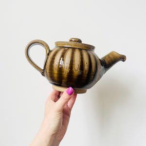 Puede incluir: Una tetera de cerámica marrón con tapa, asa y boquilla. La tetera tiene detalles acanalados verticales y una base de color claro. La tetera se sostiene sobre un fondo blanco.