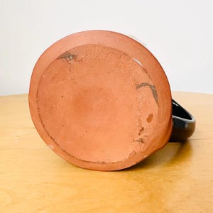 May include: A handmade ceramic mug, viewed from the bottom, showcasing a terracotta base and a black handle. The mug's base has a slightly rough texture and visible imperfections, suggesting its handcrafted nature. The mug sits on a light-colored wooden surface.