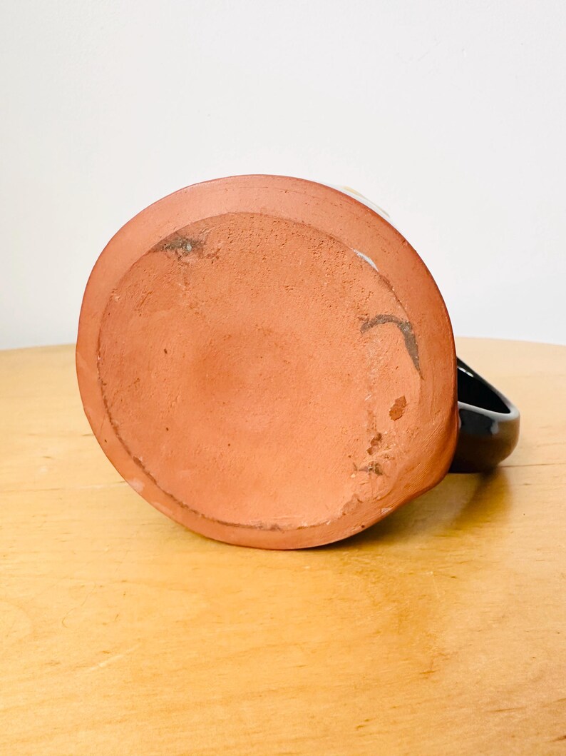 May include: A handmade ceramic mug, viewed from the bottom, showcasing a terracotta base and a black handle. The mug's base has a slightly rough texture and visible imperfections, suggesting its handcrafted nature. The mug sits on a light-colored wooden surface.