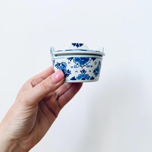 May include: A small, covered ceramic container with a blue and white floral pattern. The container has a lid with a handle and two small handles on the sides. The design features stylized flowers and leaves.