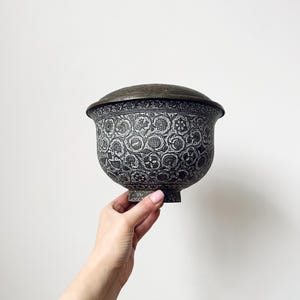 Pode incluir: Uma tigela de metal antiga com tampa, apresentando intrincados padrões florais em um esquema de cores cinza escuro e prata. A tigela tem formato arredondado com base de pedestal e tampa combinando. O design é detalhado e ornamentado, mostrando uma estética vintage.