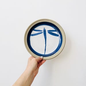 May include: A round ceramic plate with a speckled white centre and a deep blue rim. A hand-painted blue dragonfly with elongated wings and a slender body is centred on the plate. The outer edge of the plate features a natural, unglazed beige finish.