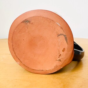May include: A handmade ceramic mug, viewed from the bottom, showcasing a reddish-brown, unglazed base. The mug has a black handle and a white interior rim. The mug sits on a light-colored wooden surface.