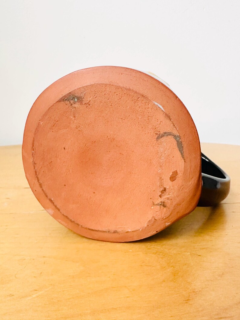 May include: A handmade ceramic mug, viewed from the bottom, showcasing a reddish-brown, unglazed base. The mug has a black handle and a white interior rim. The mug sits on a light-colored wooden surface.