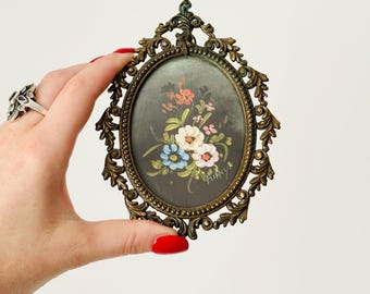 Könnte beinhalten: Ein kleiner ovaler Bilderrahmen mit einem verzierten goldenen Metalldesign. Der Rahmen enthält ein handgemaltes Blumenbild mit rosa, weißen, blauen und roten Blumen auf einem dunklen Hintergrund. Die Unterschrift des Künstlers, "Binky", ist in der rechten unteren Ecke sichtbar.