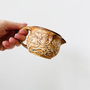 Puede incluir: Una pequeña jarra de cerámica marrón con asa y boquilla. La jarra presenta un diseño floral en relieve en un color marrón más claro. La jarra se sostiene sobre un fondo blanco.