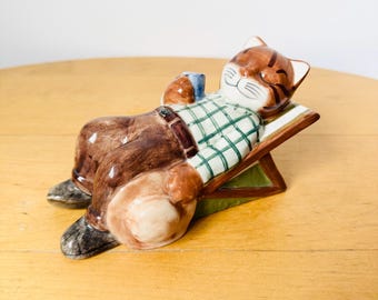 Peut inclure: Une figurine en céramique d'un chat portant une chemise à carreaux et un pantalon marron, se relaxant sur une chaise longue verte et marron. Le chat tient une tasse bleue dans sa patte.