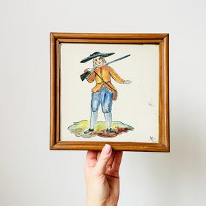 May include: A framed, square painting featuring a figure in historical attire, holding a rifle. The figure wears an orange jacket, blue pants, and a black hat. The artwork is set against a white background, framed in wood.