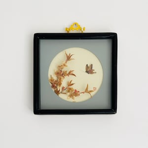 Può includere: Un'opera d'arte incorniciata con un disegno circolare di fiori secchi e una farfalla. La cornice quadrata nera ha una parte superiore decorativa gialla. Lo sfondo è grigio chiaro. Probabilmente si tratta di un pezzo decorativo.