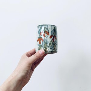 Può includere: Un bicchiere cilindrico in ceramica con base grigio chiaro e un motivo dipinto a mano di fiori arancioni e fogliame verde tenue. Lo smalto presenta una texture sottile e screziata con pennellate verticali che creano un aspetto stratificato.