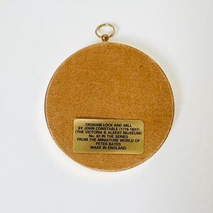 May include: A round, framed artwork with a gold-colored ring for hanging. The artwork features a tan-colored, textured surface. A rectangular gold-colored plaque with black text is affixed to the artwork, which reads "DEDHAM LOCK AND MILL" and other details.