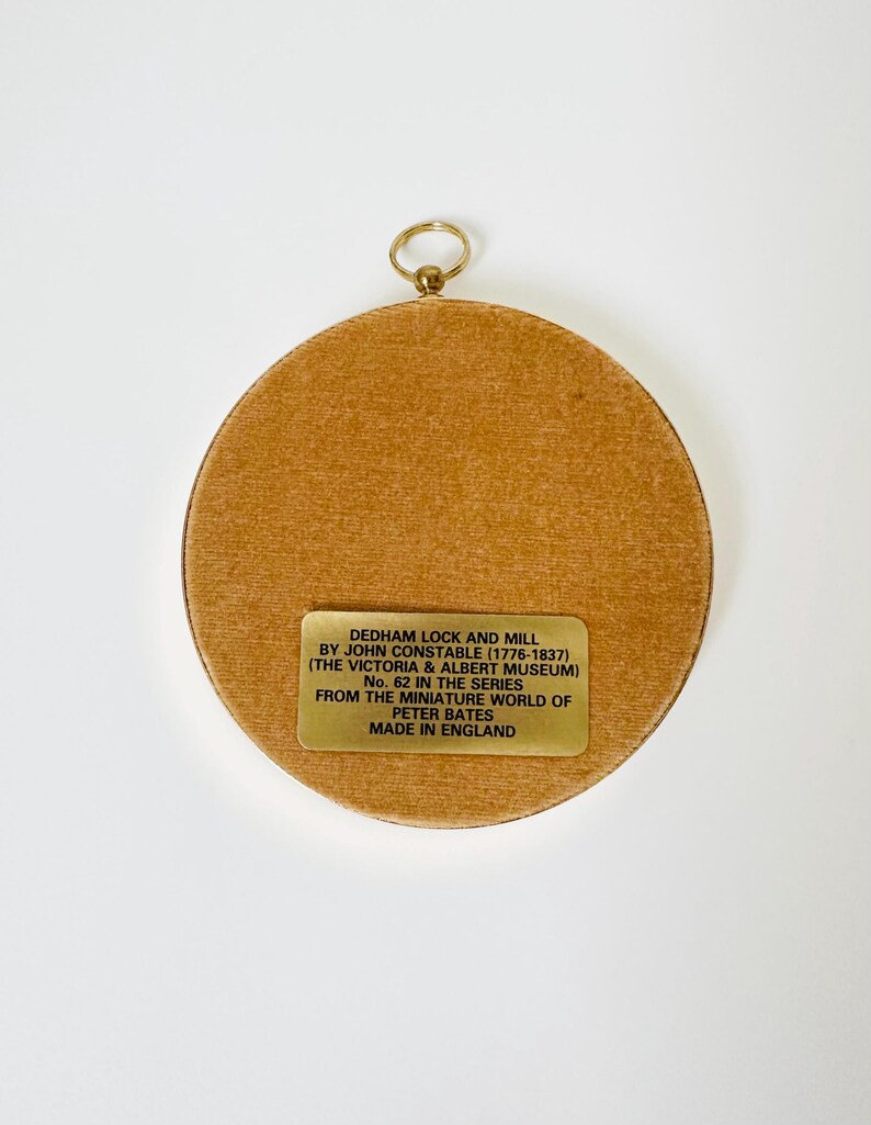 May include: A round, framed artwork with a gold-colored ring for hanging. The artwork features a tan-colored, textured surface. A rectangular gold-colored plaque with black text is affixed to the artwork, which reads "DEDHAM LOCK AND MILL" and other details.
