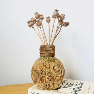 Puede incluir: Un jarrón de cerámica texturizado, en tonos marrón y verde, con cápsulas de semillas secas. El jarrón está sobre una superficie de madera, junto a un libro titulado "MAD ABOUT THE HOUSE".