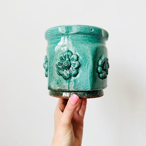May include: A turquoise ceramic planter with embossed floral designs. The cylindrical pot has a textured surface and a dark brown base. The planter is held by a hand against a white background. The pot is approximately 13 cm tall.