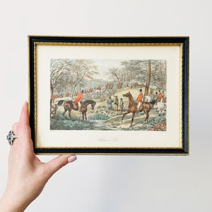 Könnte beinhalten: Gerahmter Druck, der eine Jagdszene mit Reitern auf Pferden in einem Wald darstellt. Der Druck hat einen schwarzen Rahmen mit einem goldenen Innenrand. Das Kunstwerk zeigt Figuren in roten Mänteln und eine detaillierte Landschaft.