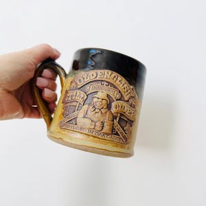 May include: A ceramic mug with a brown handle and a textured surface. The mug features a relief design with the words "Old English Cider" and a depiction of a man. The mug is tan and brown.