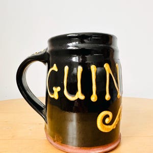 May include: Black ceramic mug with the word "Guinness" in gold lettering. The mug has a handle and a decorative swirl. The base of the mug is a light pink color.