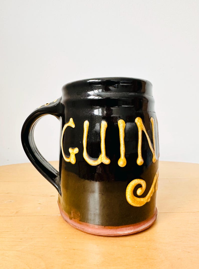 May include: Black ceramic mug with the word "Guinness" in gold lettering. The mug has a handle and a decorative swirl. The base of the mug is a light pink color.