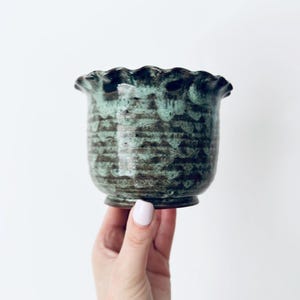 Peut inclure: Un pot en céramique avec un bord festonné et ondulé, doté d'une glaçure sombre et tachetée aux tons vert forêt profond et noir. Le récipient présente un corps arrondi qui se rétrécit vers une petite base circulaire, avec une finition texturée.