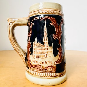 Op de afbeelding: Een bruine en blauwe keramische bierkan met een reli&euml;fbeeld van een gebouw en de tekst "Hotel de Ville".