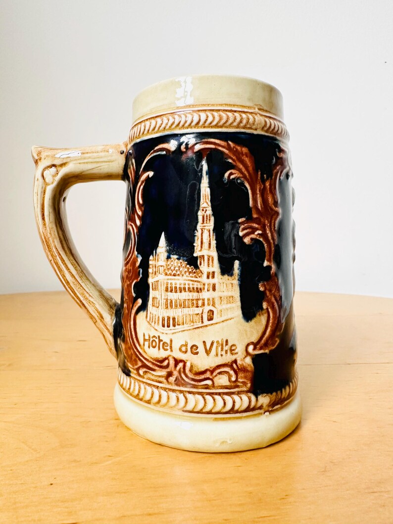 Op de afbeelding: Een bruine en blauwe keramische bierkan met een reli&euml;fbeeld van een gebouw en de tekst "Hotel de Ville".