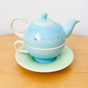 May include: A ceramic tea set for one featuring a light blue teapot with a matching lid and a cup, both decorated with small multicolored polka dots. The teapot and cup rest on a pale green saucer, creating a stacked, compact tea service.