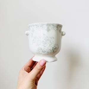 May include: White ceramic planter with a grey floral and scrollwork design. The planter has a pedestal base and two small handles. The floral pattern is repeated around the top rim. A decorative piece for home decor.