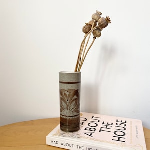 May include: A brown and white ceramic vase with a geometric design, holding dried poppy seed pods. The vase is sitting on top of a book titled "Mad About The House" by Kate Watson-Smyth.