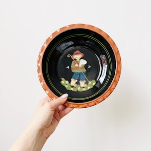 Op de afbeelding: Een ronde keramische kom met een zwart interieur en een bruine rand. De kom heeft een geschilderde illustratie van een persoon die een staf en een schaap vasthoudt. De persoon draagt een rode hoed, een groene jas en een blauwe broek.