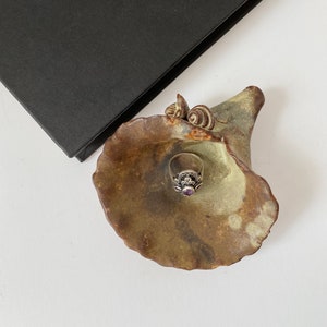 K&ouml;nnte beinhalten: Ein Keramik-Ringhalter in Form einer Blume mit einer gr&uuml;n-braunen Glasur. Ein silberner Ring mit einem violetten Stein liegt im Halter.