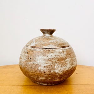 May include: A round, lidded ceramic jar with a speckled brown and white finish. The jar has a bulbous body and a small, rounded knob on the lid. The ceramic piece is sitting on a wooden surface.
