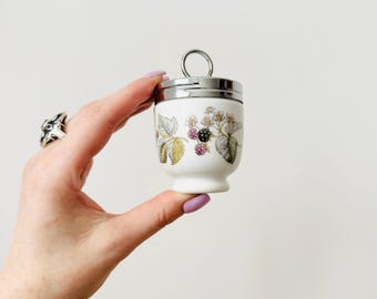 Puede incluir: Una huevera de cerámica blanca con tapa y asa plateadas. La taza está decorada con un diseño botánico de moras y hojas en tonos verdes, morados y amarillos. Un artículo de cocina clásico.