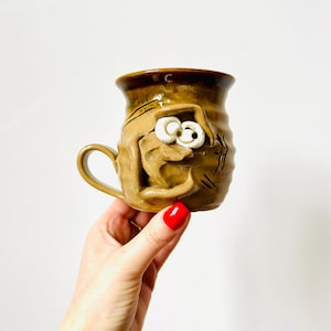 May include: A handcrafted ceramic mug with a quirky design. The mug is a warm brown colour with a sculpted face and a white eye. The handle is curved, and the mug has a glossy finish. A unique piece for coffee or tea.