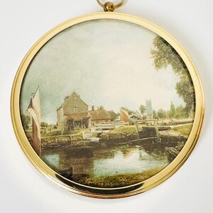 May include: A round, framed print featuring a landscape scene with a river, buildings, and trees. The print is enclosed in a gold-colored metal frame with a hanging loop at the top. The artwork depicts a serene, rural setting.