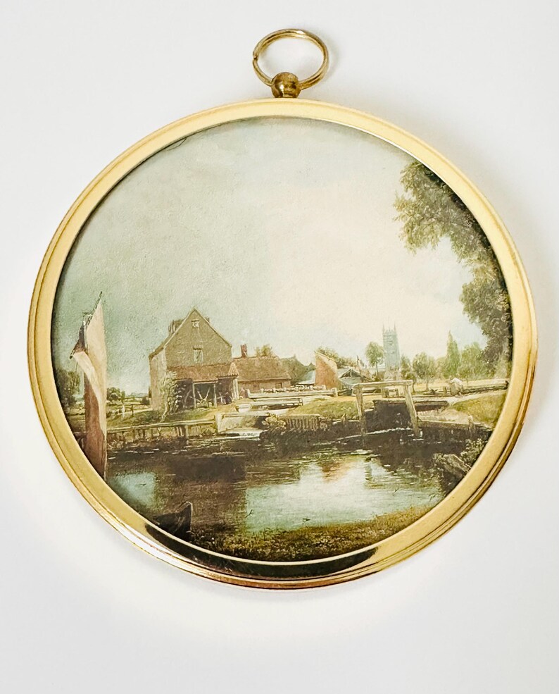 May include: A round, framed print featuring a landscape scene with a river, buildings, and trees. The print is enclosed in a gold-colored metal frame with a hanging loop at the top. The artwork depicts a serene, rural setting.