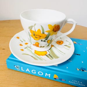 May include: White teacup and saucer set with a floral daffodil design. The cup and saucer feature yellow and white daffodils with green stems and leaves. The set is placed on a blue book with the word "LAGOM" on the cover.