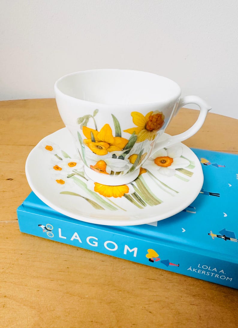 May include: White teacup and saucer set with a floral daffodil design. The cup and saucer feature yellow and white daffodils with green stems and leaves. The set is placed on a blue book with the word "LAGOM" on the cover.
