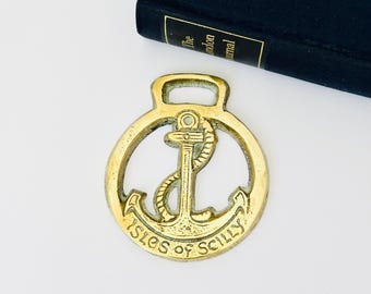 Op de afbeelding: Een cirkelvormig koperen paardenornament met een centraal ankerontwerp omwikkeld met touw. De onderrand van het cirkelvormige frame is voorzien van de tekst "ISLES of SCILLY" in reliëf. Het object heeft een kleine rechthoekige lus aan de bovenkant.
