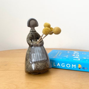 May include: A ceramic figurine of a woman in a brown dress holding a bouquet of dried yellow flowers. The figurine is standing on a wooden surface next to a blue book with the word "LAGOM" printed on the cover.