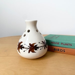 May include: White ceramic vase with a brown floral design and holes around the top. The vase is sitting on a wooden table next to two books titled "House Plants" and "Birds".