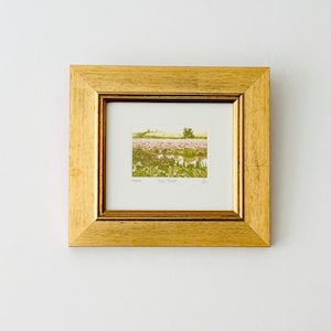 Peut inclure: Une œuvre d'art encadrée avec un cadre en bois doré. L'œuvre d'art à l'intérieur représente un paysage avec un champ de fleurs roses, un feuillage vert et un arbre sous un ciel clair. L'œuvre est encadrée avec un bord blanc.