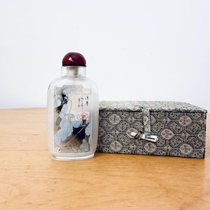Puede incluir: Un frasco de perfume de vidrio transparente con un tapón rojo oscuro y un diseño floral. El frasco está junto a una caja rectangular con un exterior de tela estampada y un cierre de cierre. El frasco tiene caracteres asiáticos.