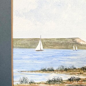 Puede incluir: Una pintura de acuarela de un paisaje mar&iacute;timo con dos veleros en la distancia. El agua es de color azul claro y el cielo es de un blanco p&aacute;lido. La costa es de color marr&oacute;n arenoso con hierba verde creciendo en ella.