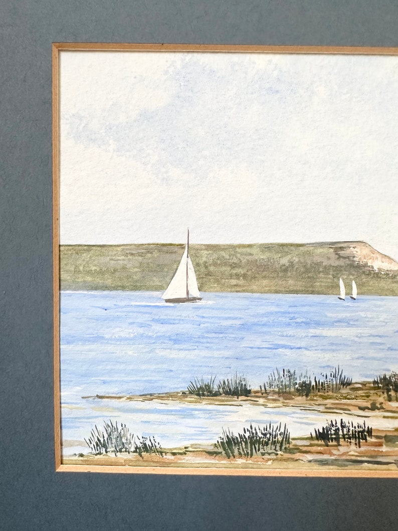 Puede incluir: Una pintura de acuarela de un paisaje mar&iacute;timo con dos veleros en la distancia. El agua es de color azul claro y el cielo es de un blanco p&aacute;lido. La costa es de color marr&oacute;n arenoso con hierba verde creciendo en ella.