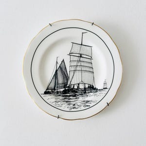 Może przedstawiać: Biały talerz ceramiczny z falistym brzegiem i złotym wykończeniem, ozdobiony czarną ilustracją przedstawiającą dwa żaglowce na wodzie. Napis J.S. Dews znajduje się w prawym dolnym rogu, a talerz jest zamocowany na czarnym drucianym uchwycie ściennym.