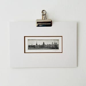 May include: A framed black and white etching of a cityscape, held by a silver clip against a white background. The artwork features buildings, spires, and ships, with fine details. The frame is a thin, warm brown.