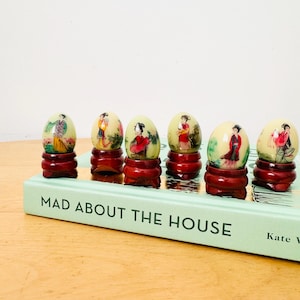 May include: Six decorative egg-shaped ornaments with painted figures on a light green background, each mounted on a small, dark red wooden stand. The eggs are arranged on a book titled "MAD ABOUT THE HOUSE".