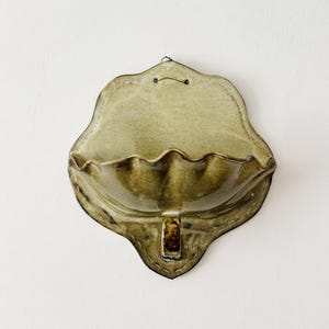 May include: A wall-mounted ceramic pocket planter featuring a glossy olive green glaze with a scalloped, shell-like basin and a flat back panel. The piece includes a small metal wire loop at the top for hanging and a decorative tab at the base.