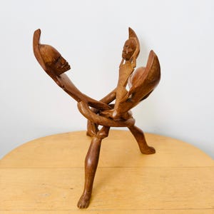 Peut inclure: Sculpture en bois sculpté représentant trois figures entrelacées. La sculpture est faite d'un bois brun chaud et présente des traits de visage détaillés et des formes fluides. Les figures sont reliées à la base, créant un sentiment d'unité.